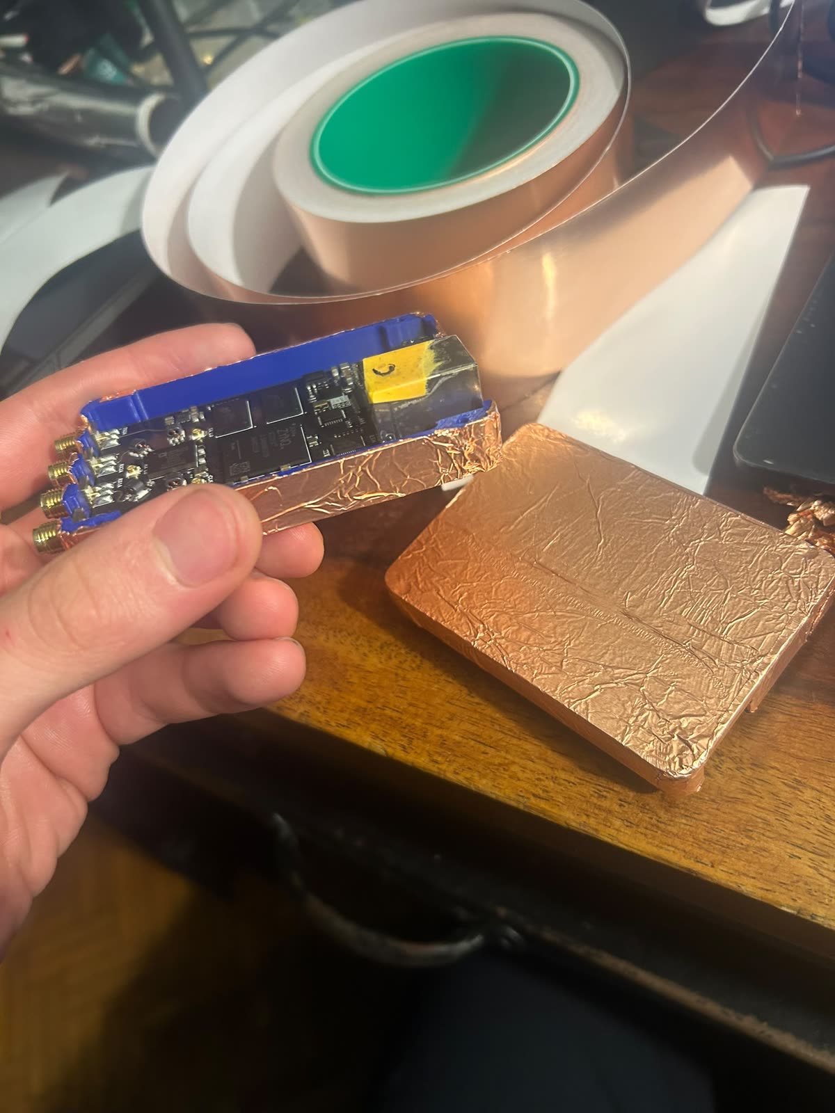 Une carte SDR bleue tenue en main, une extrémité habillée de scotch cuivre, à côté d'un rouleau de cuivre et d'un bloc blindé